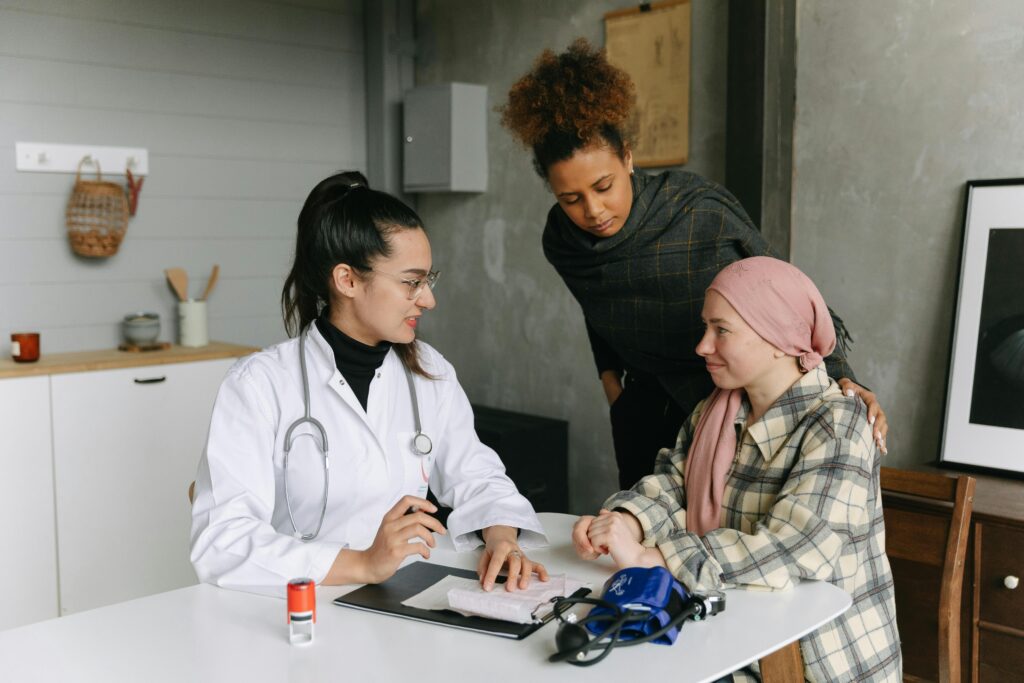 A compassionate female doctor discusses cancer remission with a young female patient wearing a headscarf, while a supportive friend stands beside her in a comforting clinical setting.